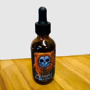 Op de afbeelding: Een bruine glazen fles met een zwarte druppelaar. Het etiket toont een blauw schedelontwerp met oranje accenten en de woorden "Wildmind Botanicals". De fles staat op een houten oppervlak.