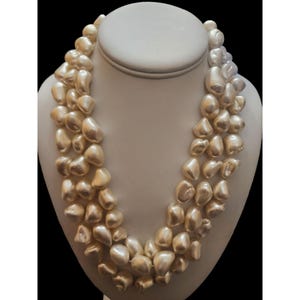 May include: A three-strand necklace featuring large, irregularly shaped, off-white pearls. The pearls have a lustrous sheen and are displayed on a white bust form against a black background. The necklace is a statement piece.
