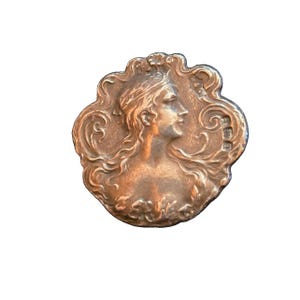 May include: A bronze-toned Art Nouveau brooch depicting a woman's profile within a decorative, swirling frame. The woman has flowing hair and a calm expression. The brooch has a detailed, textured surface and is set against a white background.