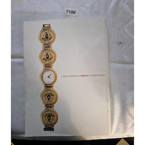 May include: A gold-coloured Versace watch with a white watch face and black hands. The watch features circular medallions with a Medusa head design. The text "IL NUOVO OROLOGIO LE MEDUSE DI GIANNI VERSACE" is printed on the page.