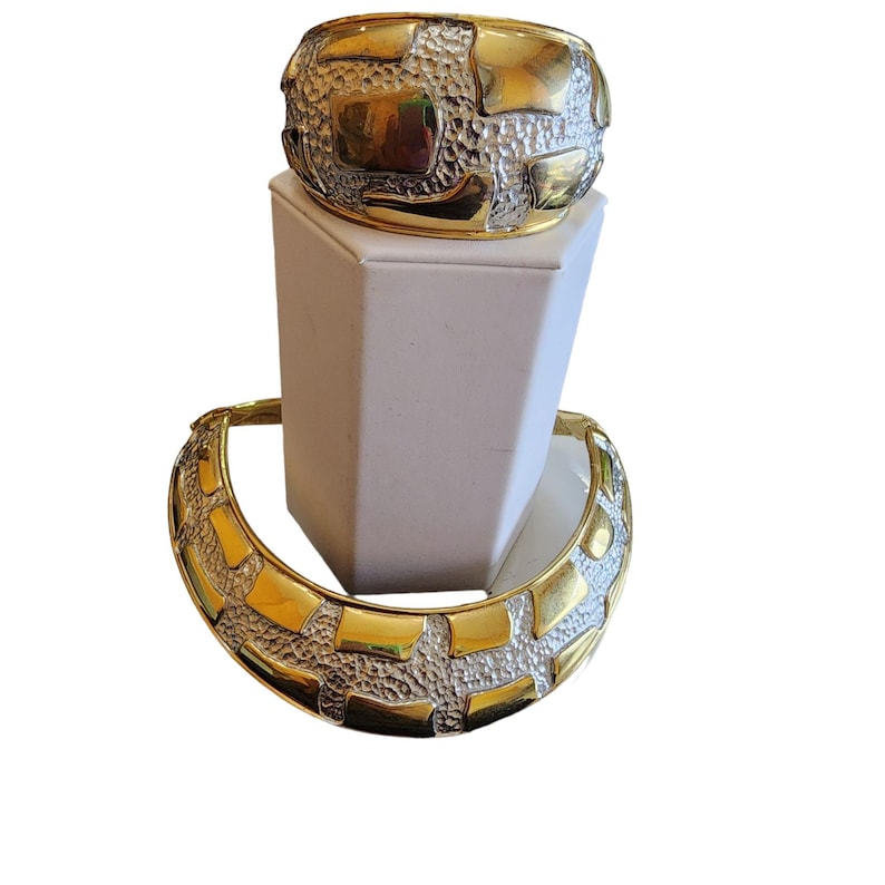May include: Two-piece jewelry set featuring a gold and silver-tone design. The set includes a wide cuff bracelet and a matching collar necklace. The cuff bracelet is displayed on a white pedestal. The jewelry has a textured, geometric pattern.