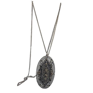 May include: A long silver chain necklace with an oval pendant featuring a filigree design. The pendant has a detailed floral pattern with a central square shape.