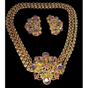 May include: A gold tone necklace with a large, ornate pendant featuring pink and purple rhinestones. The necklace is accompanied by a pair of matching earrings.