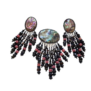 May include: A set of three pieces of jewelry: a pendant and two earrings. Each piece features an oval abalone shell with a beaded fringe in black, pink, and iridescent beads. The jewelry is set against a white background.
