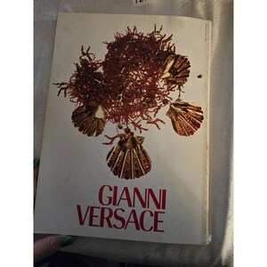 May include: A vintage book cover featuring the name "GIANNI VERSACE" in red lettering. The cover design includes a cluster of reddish-brown coral and gold seashell ornaments against a cream-colored background. The book appears to be a fashion or design publication.
