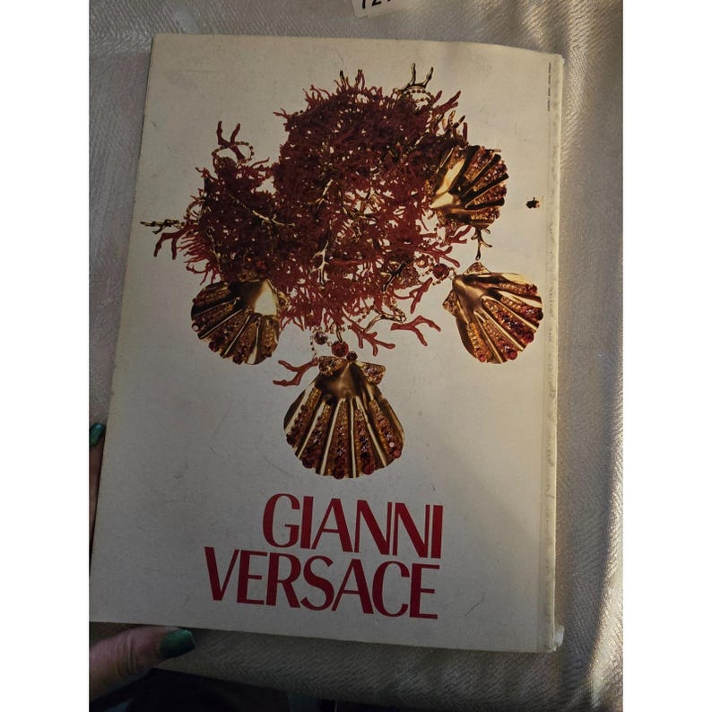 May include: A vintage book cover featuring the name "GIANNI VERSACE" in red lettering. The cover design includes a cluster of reddish-brown coral and gold seashell ornaments against a cream-colored background. The book appears to be a fashion or design publication.