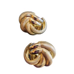 May include: Two gold-toned, textured clip-on earrings. Each earring is a knot design with a ribbed texture and a polished, smooth finish on the knot's center. The earrings are set against a white background.