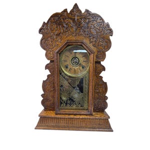 May include: An antique wooden clock with intricate floral carvings. The clock face is gold-toned with black numerals and hands. The clock has a rectangular shape with a decorative top and base. The clock's dimensions are approximately 12 inches wide, 20 inches tall, and 6 inches deep.