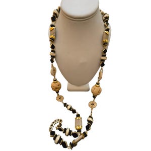 May include: A long necklace with a variety of beads in black, cream, and gold tones. The necklace features geometric and organic shapes, including diamond-shaped beads, round beads, and textured, spherical beads. The necklace is displayed on a white mannequin.