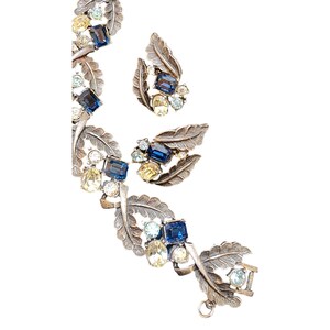 Op de afbeelding: Een zilverkleurige armband en oorbellen set met blauwe en gele strass stenen in een bladontwerp.