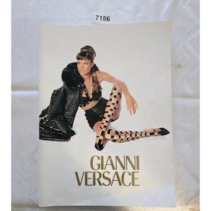 May include: A fashion advertisement featuring a woman in a black outfit and knee-high boots with gold accents. The text "GIANNI VERSACE" is displayed in gold lettering at the bottom of the image.