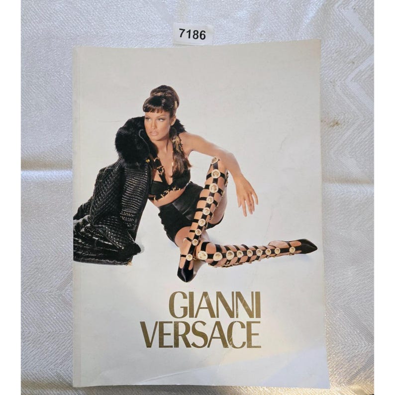 May include: A fashion advertisement featuring a woman in a black outfit and knee-high boots with gold accents. The text "GIANNI VERSACE" is displayed in gold lettering at the bottom of the image.