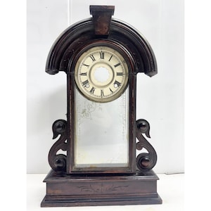 May include: Antique wooden mantel clock with a dark brown finish. The clock features a round face with Roman numerals, a mirrored panel, and decorative scrollwork. The clock sits on a rectangular base and has an arched top.
