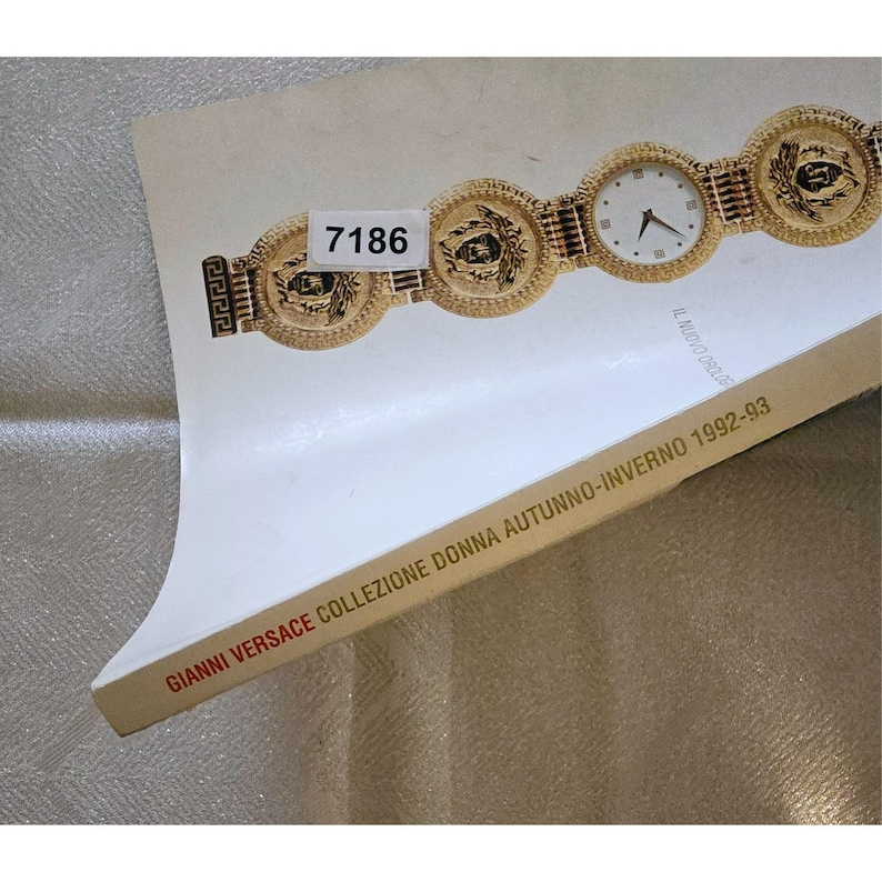 May include: A gold wristwatch with a white watch face and circular medallions. The watch is displayed on a white surface with the text "GIANNI VERSACE COLLEZIONE DONNA AUTUNNO-INVERNO 1992-93". A sticker with the number "7186" is also visible.