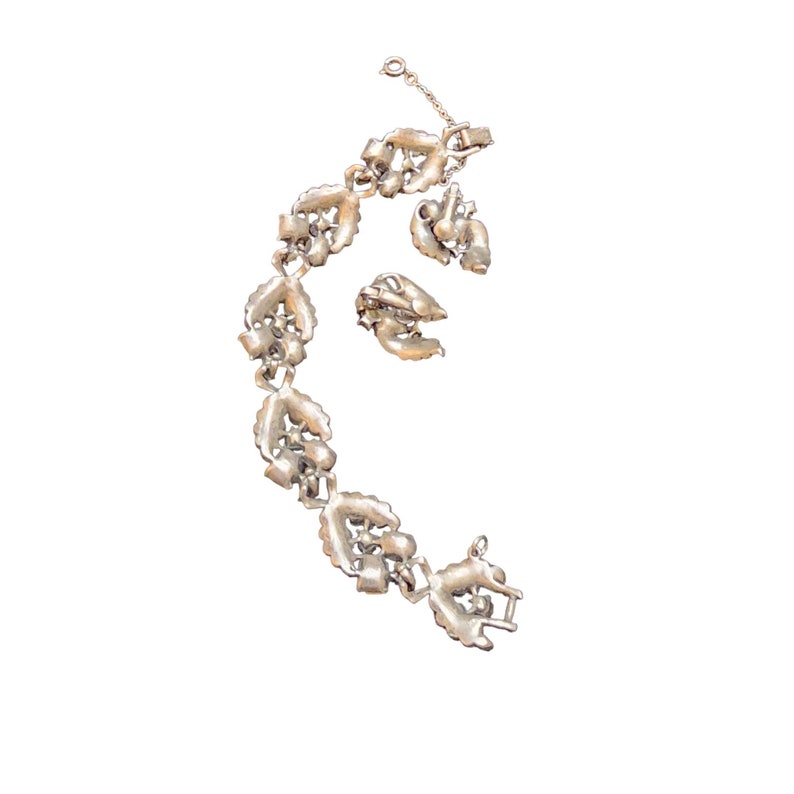 Op de afbeelding: Een zilveren armband met een hartvormig ontwerp en heldere strass steentjes. De armband heeft een sluiting en wordt geleverd met twee bijpassende oorbellen.