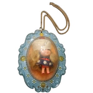 May include: A vintage pendant necklace featuring a small doll encased in a clear plastic bubble. The doll has blonde hair and wears a blue and red outfit. The pendant is set in a light blue frame with decorative accents and hangs from a gold-colored chain.