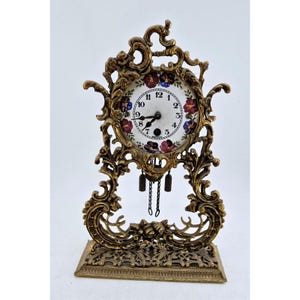 May include: An antique brass-colored mantel clock with a floral design on the clock face. The clock has a decorative, ornate frame and a rectangular base. The clock face features black numerals and hands, with a floral border.