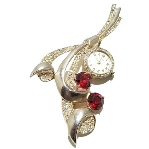 Könnte beinhalten: Eine silberne Brosche mit einem Zifferblatt und roten Strasssteinen. Die Brosche ist in Form einer stilisierten Blume mit einem langen Stiel gestaltet.