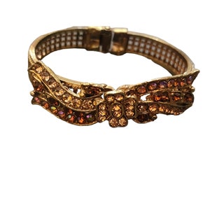 May include: A gold-tone bangle bracelet with a decorative design of a bow made of clear and amber-colored rhinestones.