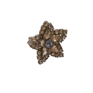 Puede incluir: Un anillo en forma de estrella con una banda plateada. La estrella está incrustada con pedrería transparente y ahumada de varios tamaños, con una sola pedrería azul claro en el centro. El anillo está sobre un fondo blanco.