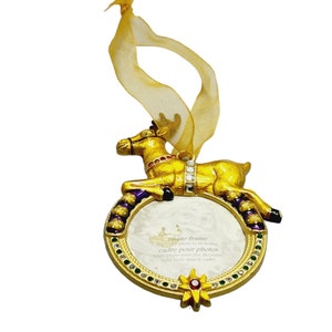 May include: Gold-tone reindeer ornament with a white photo frame. The frame has a gold rim with green and purple enamel accents and rhinestones. The reindeer is sitting on the frame and has red, green, and white enamel accents. The ornament has a gold ribbon for hanging.