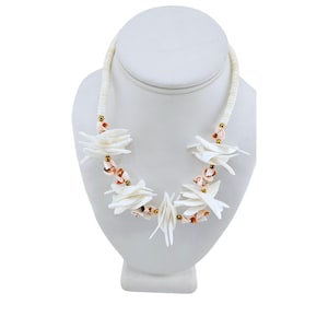 May include: A white shell necklace with a unique design. The necklace features white shell flowers and small, round gold-colored beads. The necklace is displayed on a white mannequin head.