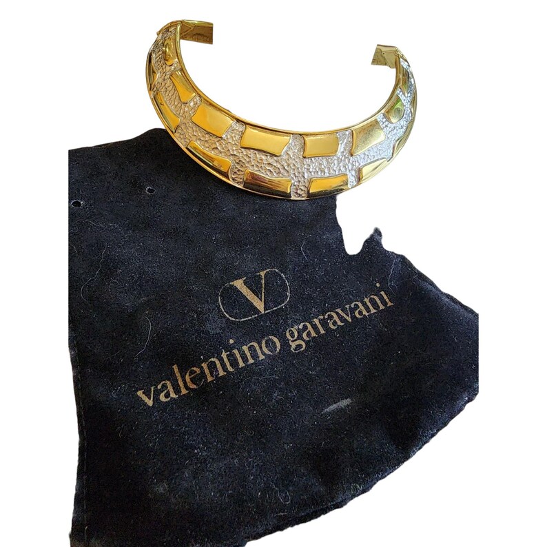 May include: A gold-toned cuff bracelet with a textured, geometric design. The bracelet is displayed on a black velvet pouch with the text "valentino garavani" in gold lettering.