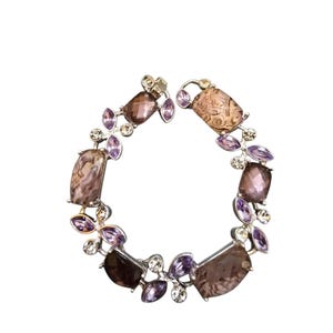 Könnte beinhalten: Ein silbernes Armband mit einer Reihe von braunen und lila rechteckigen und ovalen Steinen. Das Armband ist mit kleineren, blattförmigen lila Steinen und klaren Kristallen verziert, wodurch ein funkelndes, elegantes Design entsteht. Ein wunderschönes Schmuckstück.