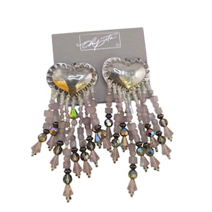 May include: Silver heart-shaped earrings with beaded fringe. The earrings have a silver heart-shaped top with a beaded fringe in shades of pink, gray, and iridescent beads. The earrings are attached to a gray card with the brand name "Chipeta".