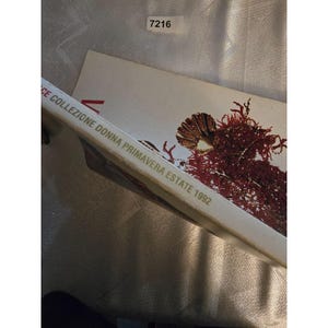 May include: A book titled "CE COLLEZIONE DONNA PRIMAVERA ESTATE 1992" with a gold and red illustration. The book is on a textured surface. A small white sticker with the number "7216" is in the upper right corner.