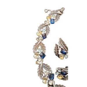 Op de afbeelding: Een zilverkleurige armband en oorbellen set met blauwe, gele en heldere strass stenen in een bladontwerp.