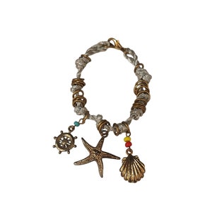 May include: A silver and gold bracelet with a nautical theme. The bracelet features a silver cord with gold rings and three charms: a gold starfish, a gold seashell, and a gold ship's wheel.