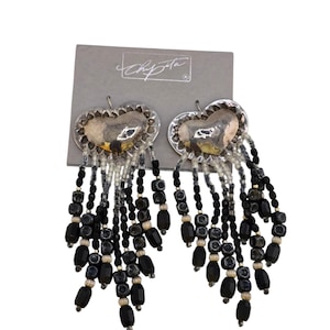 May include: Heart-shaped silver-tone earrings with dangling black and clear beads. The earrings have a textured silver heart design and are attached to a gray card with the designer's signature.