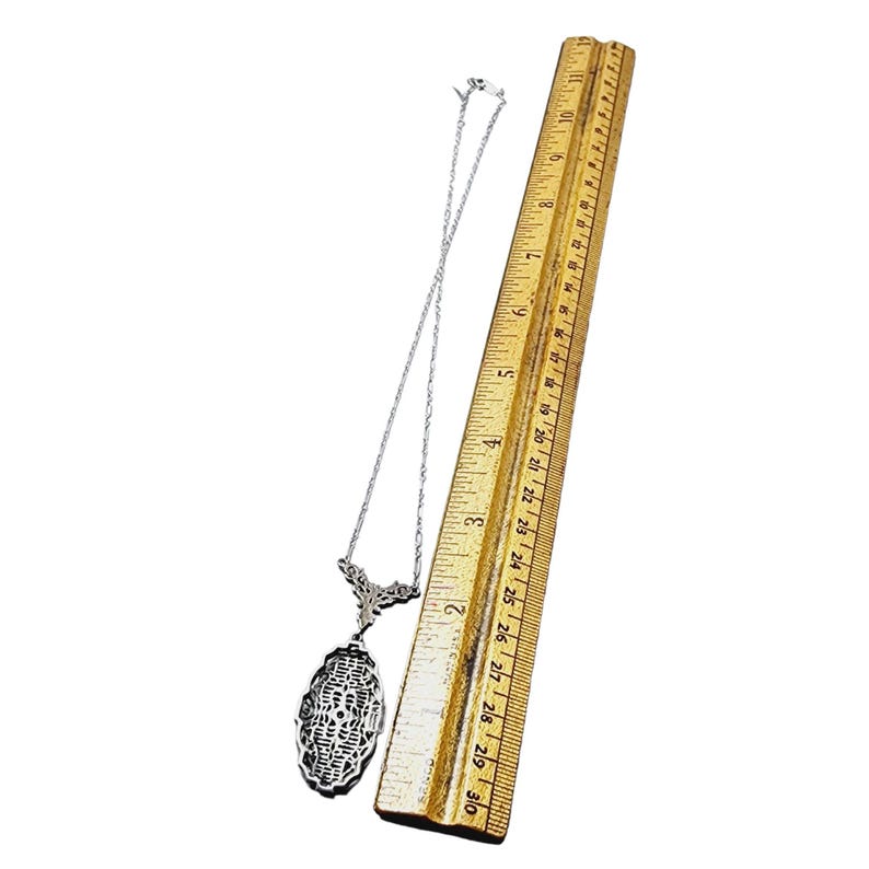 Puede incluir: Collar con colgante de filigrana plateada con dise&ntilde;o ovalado, expuesto junto a una regla de madera. El colgante mide aproximadamente 5 cm de largo. La cadena del collar es delicada y el colgante tiene un intrincado dise&ntilde;o calado.