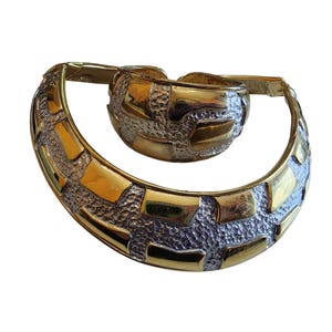 May include: A gold-toned metal jewelry set featuring a textured design. The set includes a wide, curved collar necklace and a matching cuff bracelet. The pieces have a geometric pattern with gold and silver-toned accents.
