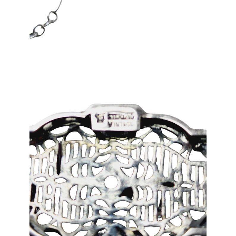 Puede incluir: Primer plano de un colgante vintage de filigrana de plata de ley. El colgante tiene un dise&ntilde;o calado con patrones intrincados y un peque&ntilde;o agujero redondo. El texto "STERLING VINTAGE" est&aacute; estampado en la parte superior. Se adjunta una delicada cadena de plata.