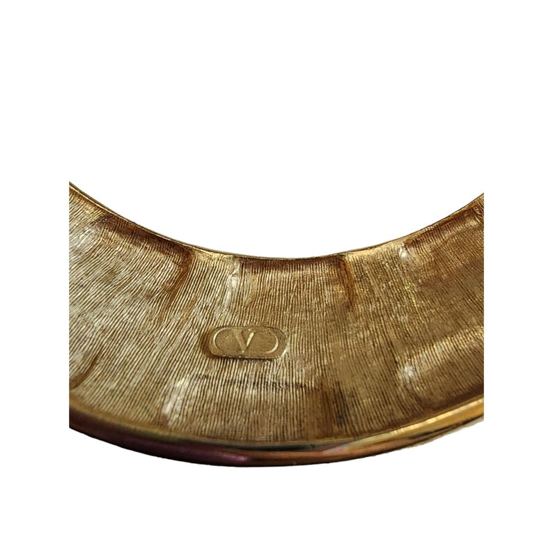 May include: Close-up of a gold-toned bracelet with a textured surface. The bracelet features the Valentino logo. The design has a ridged pattern, and the overall aesthetic is vintage.
