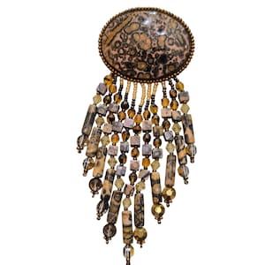 May include: An oval-shaped brooch with a speckled, light brown and black center, framed by a gold-toned metal. The brooch features dangling strands of beads in shades of brown, amber, and clear, creating a layered, fringed effect. The beads are various shapes and sizes.