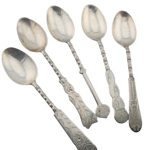 Puede incluir: Conjunto de cinco cucharas antiguas de plata con mangos ornamentados. Cada cuchara presenta un diseño de mango diferente, incluyendo motivos de bambú, florales y de monedas. Las cucharas están dispuestas sobre un fondo blanco, mostrando sus intrincados detalles.