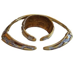 May include: A gold-toned metal cuff bracelet and two matching hinged bracelets. The bracelets feature a textured silver-colored design on the outer edges. The cuff bracelet is a wider band, while the hinged bracelets are more slender.