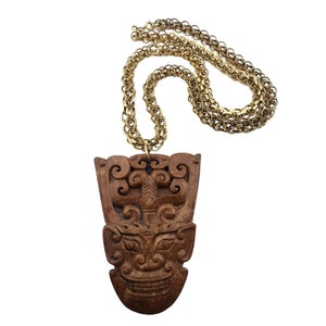 Op de afbeelding: Een goudkleurige ketting met een gesneden bruine hanger. De hanger heeft een gedetailleerd gezichtsontwerp met ingewikkelde patronen en een glimlachende uitdrukking. De ketting is gemaakt van kleine, cirkelvormige schakels.