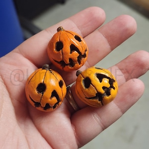 May include: Three miniature orange pumpkin figurines with carved jack-o'-lantern faces. The pumpkins have dark brown stems and are held in a hand. The pumpkins are a bright orange color with black carved features.