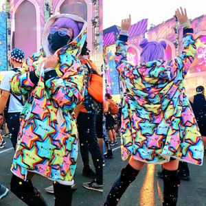 May include: A vibrant hooded coat featuring a star design in blue, yellow, pink, and green. The coat has a white, fur-lined hood and cuffs. The person is wearing black boots and a black face mask.