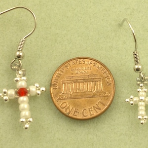May include: A pair of silver earrings with white bead cross charms and a red bead in the centre. The earrings are shown next to a UK penny for size comparison.