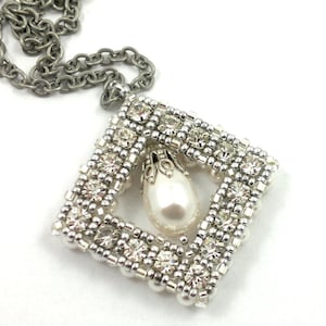 Op de afbeelding: Zilverkleurige ketting met een vierkante hanger. De hanger heeft een kralenrand, heldere kristallen en een centrale peervormige imitatieparel. De ketting heeft een schakelontwerp.