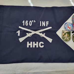May include: Navy blue flag with white text and crossed rifles. The text reads "160th INF 1 HHC". Several military patches are to the right of the flag. The patches have various designs and colors, including a lion and a number 6.