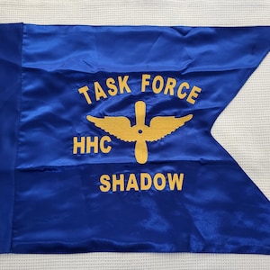 May include: A blue satin flag with the text "TASK FORCE HHC SHADOW" in gold lettering. A gold propeller and wing design is centered on the flag. The flag has a triangular end.