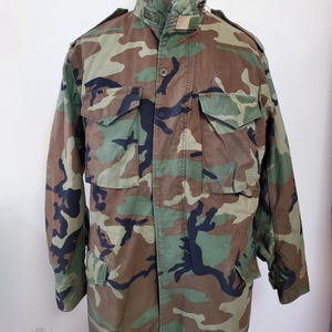 May include: Camouflage pattern military jacket with a high collar, multiple front pockets, and button and zipper closures. The jacket features a woodland camouflage design in shades of green, brown, and black.