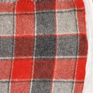 Op de afbeelding: Een close-up van een rood, grijs en zwart geruite deken met franjes. De deken heeft een klassiek vierkant patroon met een mix van rode, grijze en zwarte vierkanten. De franjes zijn rood, grijs en zwart.