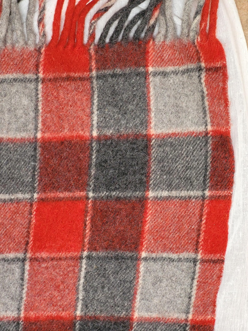 Op de afbeelding: Een close-up van een rood, grijs en zwart geruite deken met franjes. De deken heeft een klassiek vierkant patroon met een mix van rode, grijze en zwarte vierkanten. De franjes zijn rood, grijs en zwart.